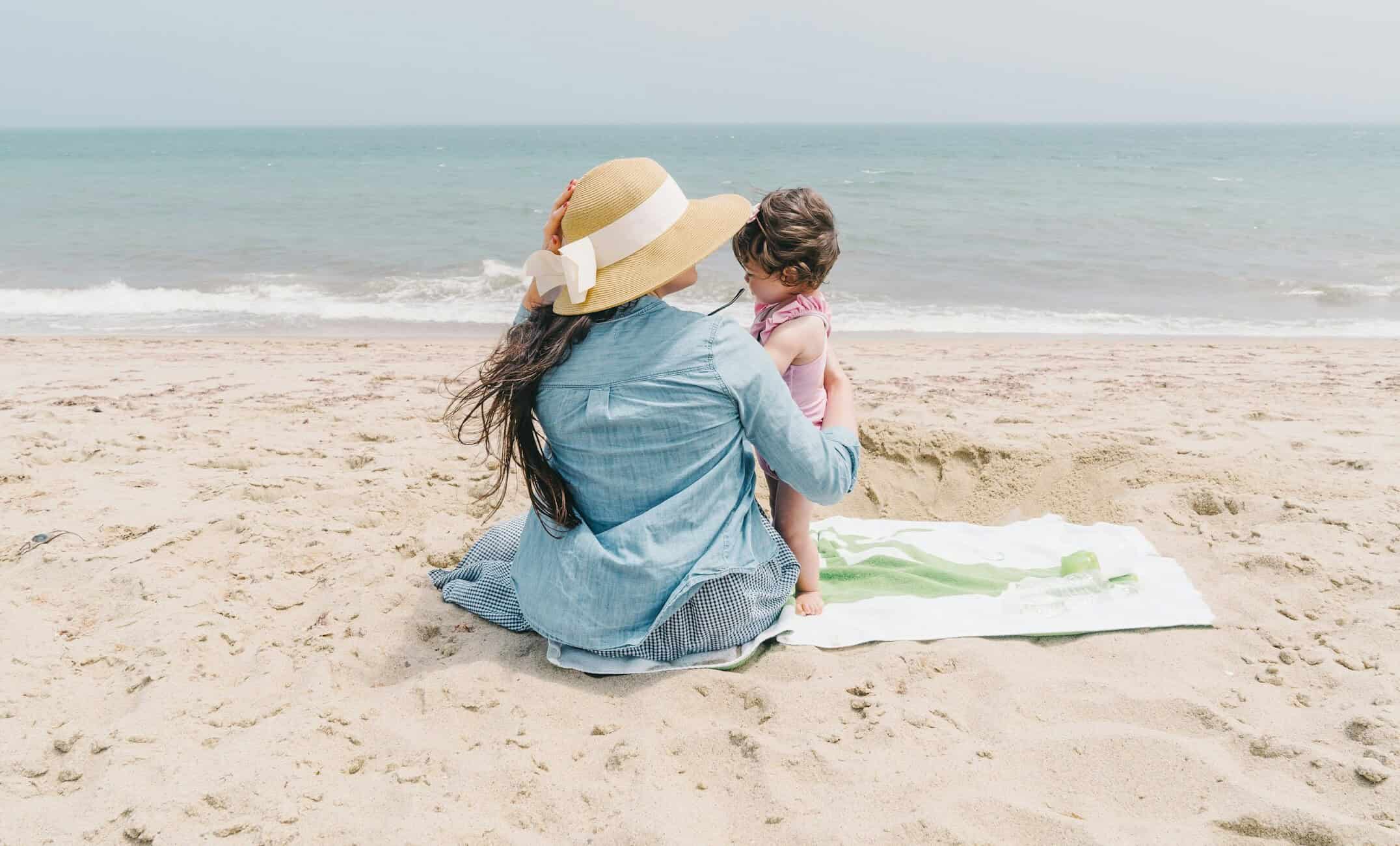 Imaginea ilustrează o mamă și copilul ei așezați pe o pătură de plajă, privind spre mare, pe o zi însorită.