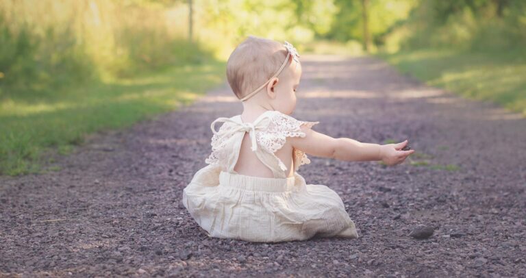 Un bebeluș pe o cărare de pietriș, îmbrăcat într-o rochie deschisă, întinzând mâna spre pământ. Fundal verde și luminos.